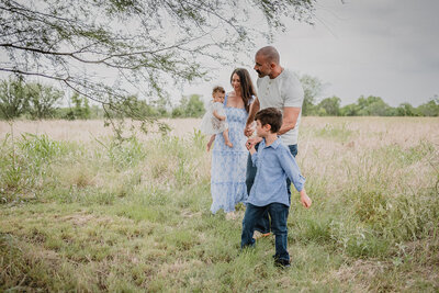 Family of four walking in field at sunset with young son leading the way during lifestyle session in New Braunfels, Texas.