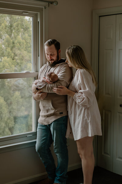 Dad in tan and jeans while standing next to mom in pink while holding baby in nursery in their home in Severn Maryland photographed by Bethany Simms Photography