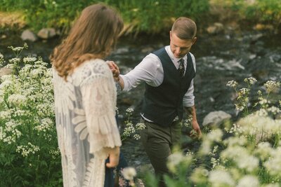 groom leads bride to a river bed amongst tall aildflowers