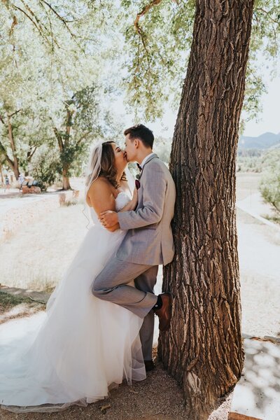 Bride kisses groom leaning against tree