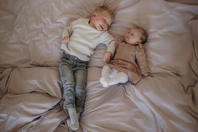Two siblings lay close to each other on a bed, smiling at each other.