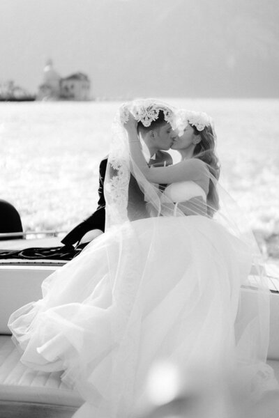 Black and white photo of bride and groom kissing under lace veil on a boat, romantic and cinematic wedding moment