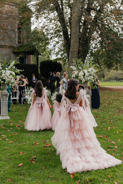 Three young girls in tiered blush pink dresses with oversized bows walk across the lawn at a garden wedding ceremony.