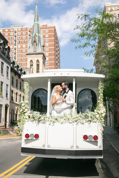 A newlywed couple dressed in a wedding gown and suit are sharing a moment on the back of a decorated white vehicle, surrounded by white flowers, traveling through a city street with a church and buildings in the background on a sunny day.