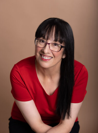 Middle aged brunette author headshot with red shirt smiling with taupe studio backdrop.