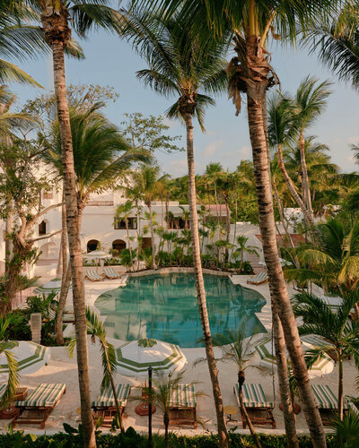 A serene resort scene featuring a hexagonal pool surrounded by palm trees. Lounge chairs with striped cushions and umbrellas are set around the pool. The resort buildings are white with arches, and the sky is clear and blue.