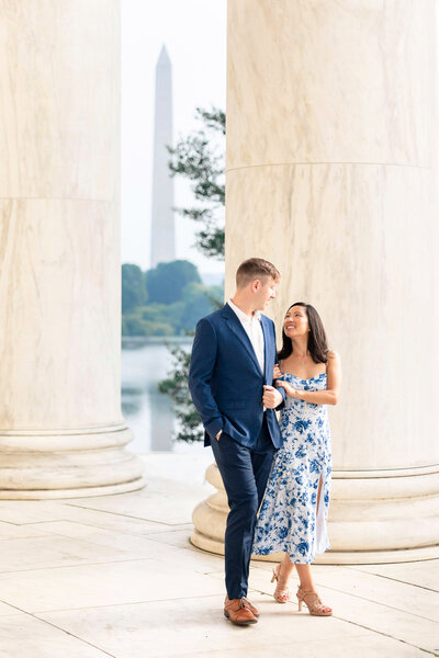 A couple walks together, smiling, on the steps of a marble building. The woman wears a white and blue floral dress, while the man is in a blue suit. The Washington Monument is visible in the background.