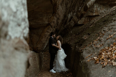 bride in lace gown and groom in dark suit laughing in an embrace beneath rocks during an adventurous elopement