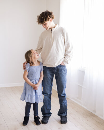 Brother and young sister standing in studio room by a window looking at each other smiling.  Sister has short blonde hair, wearing a purple dress, and black leggings.  Brother is wearing a light tan long sleeve shirt and blue jeans with short brown hair.