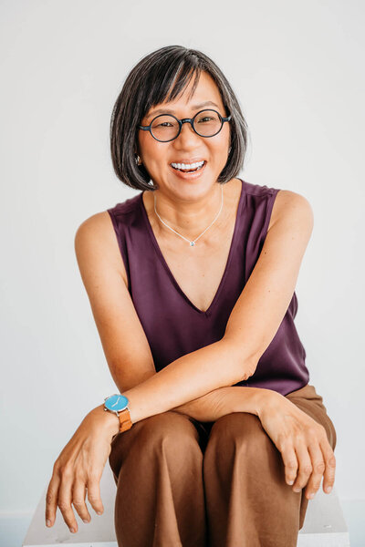 Portrait of a smiling woman with short hair, wearing a purple sleeveless top, sitting with hands clasped.