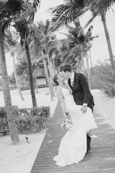 A groom dips his bride and kisses her on a pathway lined with palm trees in Cancun