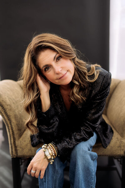 A woman with brown hair sits in a tan armchair, smiling softly and reflecting calm confidence, creativity, and warmth.