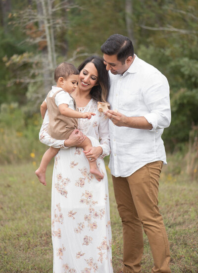Mom and dad smile while holding their toddler in an open field. A heartfelt outdoor family session by Moments in Light Photography.