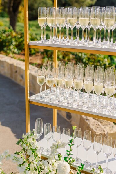 Champagne flutes filled with prosecco on a golden bookcase decorated with florals, ready to welcome guests to the wedding ceremony at Monserate Winery's Tuscan Estate in Fallbrook.