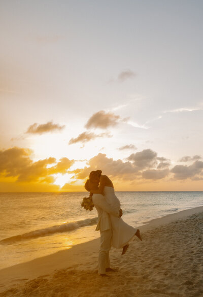 Groom lifts his bride off the sand during golden hour on the Cancun shoreline, with waves and sunset in the background.