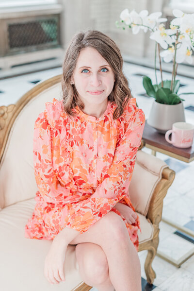 Woman looking at the camera sitting on a cram couch wearing a patterned dress