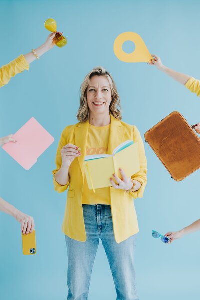 Woman with shoulder-length blonde hair wears a yellow blazer and jeans while holding a book in front of an aqua backdrop.Several other hands frame the photo holding multiple objects including sunglasses, an iPhone, a suitcase, and more.