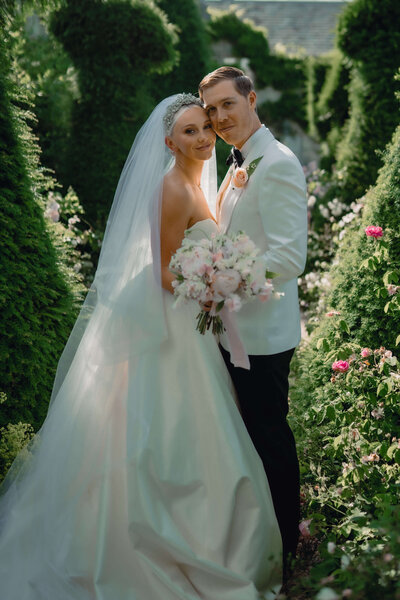 bride and groom pose together in the middled of a sculpted garden at their wedding venue euridge manor the bride holds a large pink and white bouquet