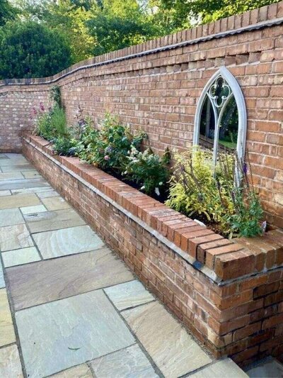 Red brick garden wall with raised flower beds and arched stone insets, bordered by sandstone paving and surrounded by trees.