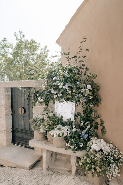 Wedding welcome signage attached to a garden wall outside the cliffside ceremony. Signage is surrounded by florals and greenery climbing up the wall and bundles of flowers filling casual wrapped baskets.