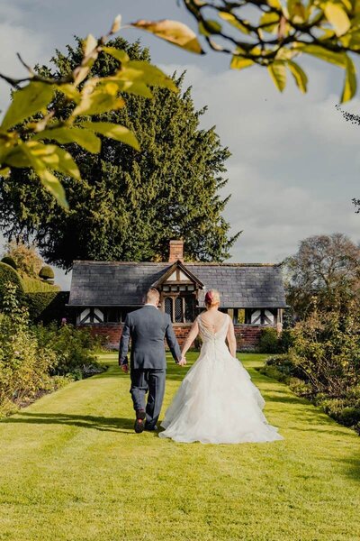 A bride and groom walking hand-in-hand down a gravel path, surrounded by lush greenery, captured candidly by a UK wedding photographer.

