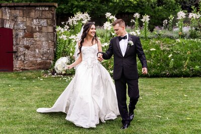 A newlywed couple walks hand in hand across a manicured lawn in Lancaster, smiling at each other in front of a blooming garden of white lilies and a rustic stone structure. The bride’s flowing satin gown features delicate embroidery, and she carries a white rose bouquet with baby’s breath, while the groom wears a classic black tuxedo with a boutonnière.