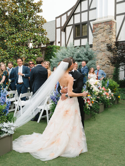 Bride and groom kissing at the end of the aisle while she holds her bouquet and her veil blows in the wind