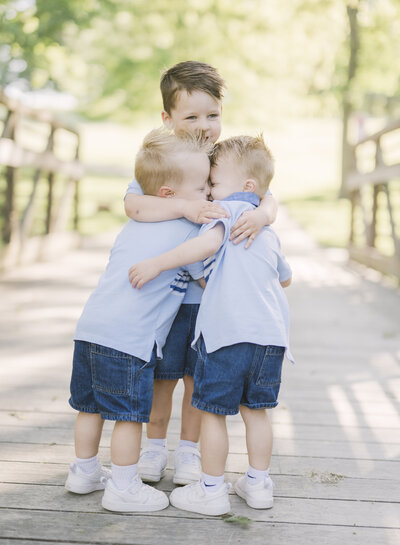 Three young boys in matching blue outfits hugging on a wooden bridge during an outdoor family session with a Kansas City family photographer.