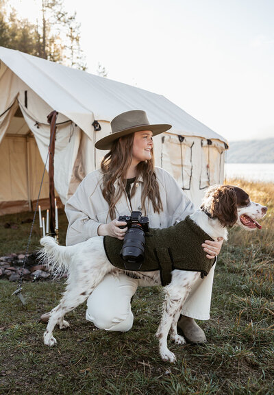 Sydney Breann, a Montana-based elopement photographer, kneels beside her dog wearing a cozy vest in front of a canvas tent at a lakeside campsite, holding her camera and smiling.