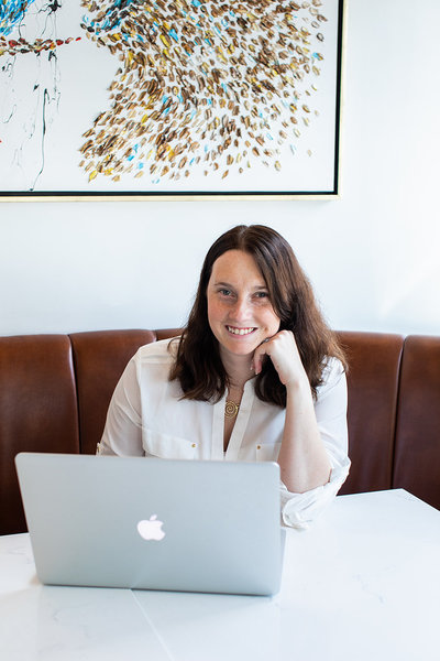 Catherine sits smiling at a white table with a silver MacBook in front of her, wearing a white button-down shirt and a gold necklace. Behind her is a modern art piece and a brown leather banquette, capturing a relaxed yet professional vibe that reflects the calm clarity her systems bring.