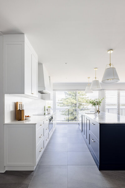 Modern kitchen with white cabinets, a navy island, and pendant lights, enhanced by large windows overlooking lush greenery—a testament to expert craftsmanship by a Newcastle interior designer.