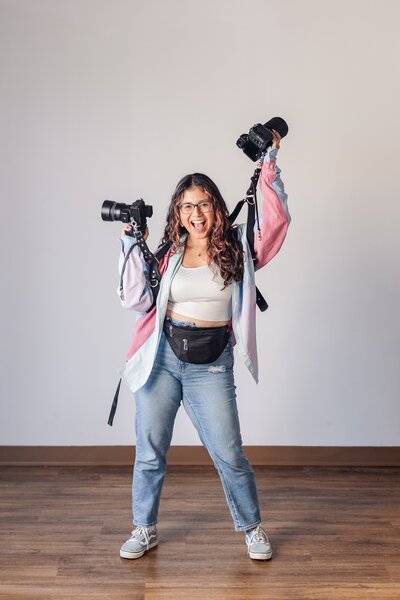 latina woman photographer dances in a colorful button up shirt with two cameras on a leather camera harness and smiles widely