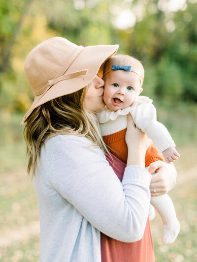 Side view of a mother wearing a floppy hat giving her six month old daughter a kiss on the cheek as the baby smiles at the camera.