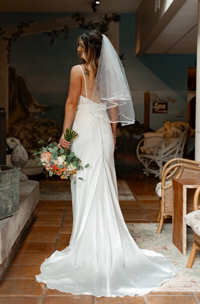 A bride stands with her back to the camera, adorned in a white wedding dress with a long train and veil. She holds a bouquet of flowers against a backdrop of wicker furniture and a mural. The scene is captured with the elegance typical of fine art weddings in New England.