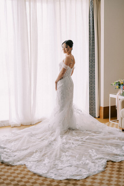 Bride standing in lace gown with long train by window, captured in natural light by Toronto photographer