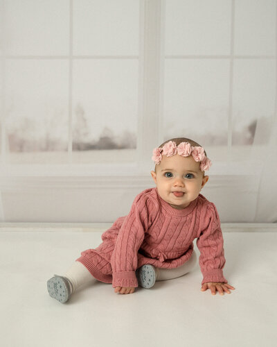 Baby girl in pink sweater dress and pink flower headband with tongue sticking out. Sitting in front of a window background.