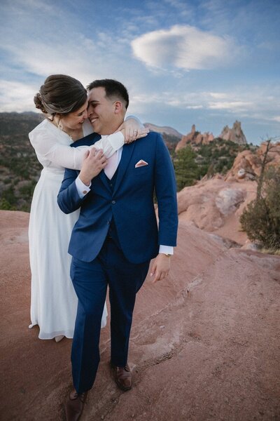 A bride stands behind her groom, wrapping her arm around his chest and he looks back at her with faces close.