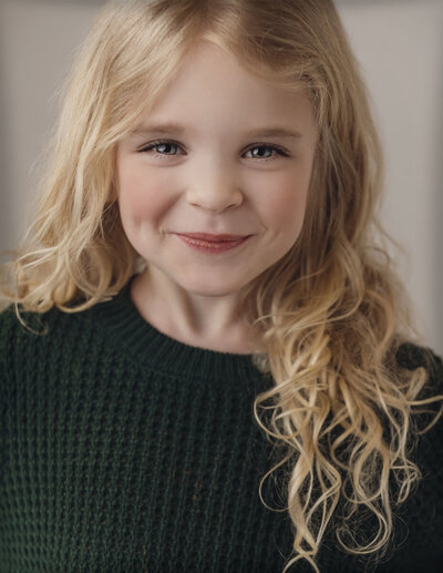 Young child with long blonde hair smiling softly, wearing a green knit sweater. The background is softly blurred, creating a warm, cheerful tone.