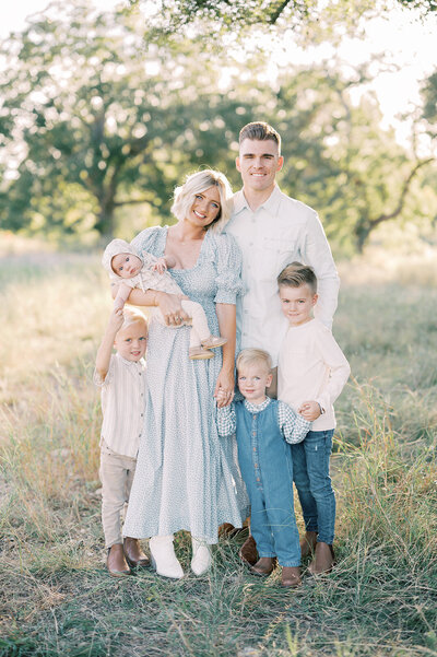 a beautiful family of 5 stands in a field during golden hour light, photo taken by San Antonio Newborn Photographer Teressa Jane