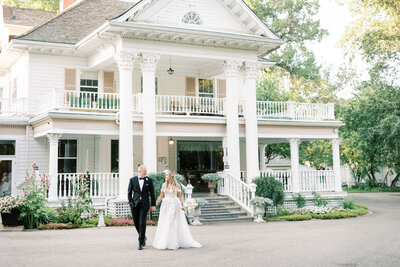 A bride and groom walk arm in arm in front of a large white, two-story house with pillars, balconies, and ornate detailing. The exquisite wedding design is surrounded by lush greenery and flowers.