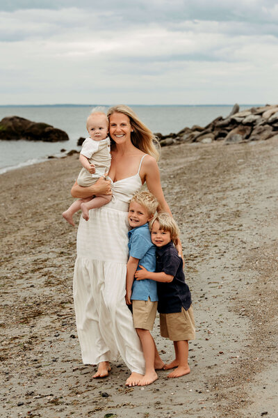 Family of five standing together on a rocky beach, with parents holding their three young children against a backdrop of calm ocean waters and overcast sky