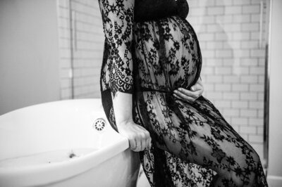 photo of a pregnant woman wearing a black lace dress sitting on the edge of a tub with her hand on her belly