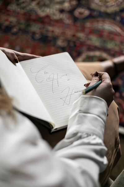 Ashleigh C. Henry, life coach and author, writing in a journal with the name of her hospitality brand, The House of Harmony, near a vintage red and maroon rug, in neutral trousers, white blouse, at the Blind Tiger Asheville, NC. 