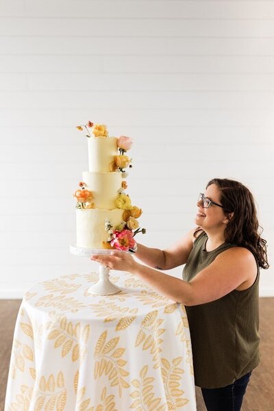 A girl with brown hair and a green shirt adjusts a wedding cake on a table. The cake is three tiers with yellow frosting and fresh flowers.