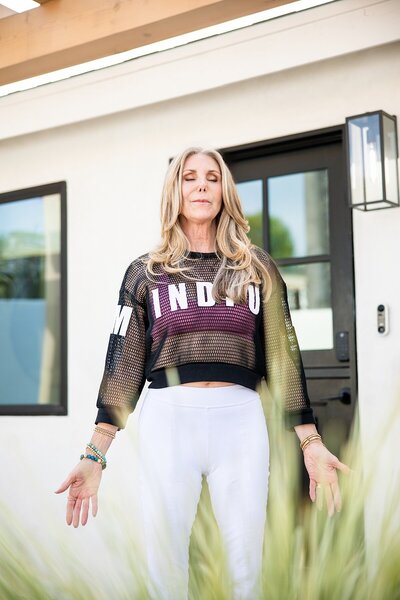 Woman in a black mesh “MINDFUL” top and white leggings stands with eyes closed and hands relaxed at her sides, practicing mindfulness in front of a modern home. Captured during a personal branding photoshoot, this image reflects calm, alignment, and holistic wellness branding.