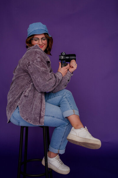 Rachel Hahn, owner, smiles with her camera in hand in front of a purple backdrop wearing denim.