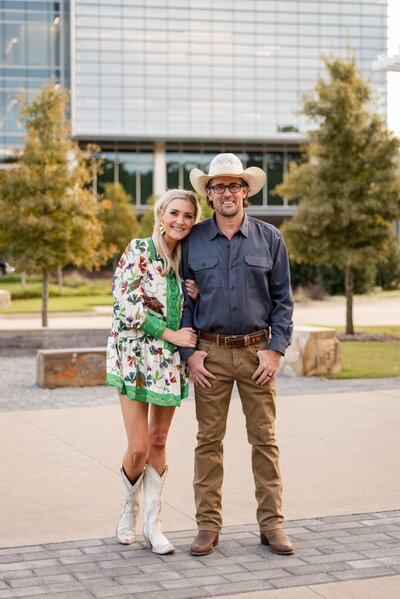 David Munsey III and his wife standing close together and smiling in front of a modern building, dressed in western-inspired outfits.
