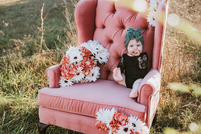 Six month milestone photo of baby girl in black with green bow sitting in pink chair holding her foot in a field in Annapolis Maryland.