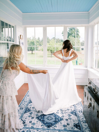 A bridesmaid helps a bride with her wedding dress train, representing moments cherished together at Oregon weddings when hiring Birdie June event planners. 