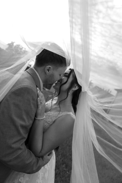 Bride and groom sharing a kiss under the wedding veil, captured in a tender and intimate moment, with the veil gently draped over them.